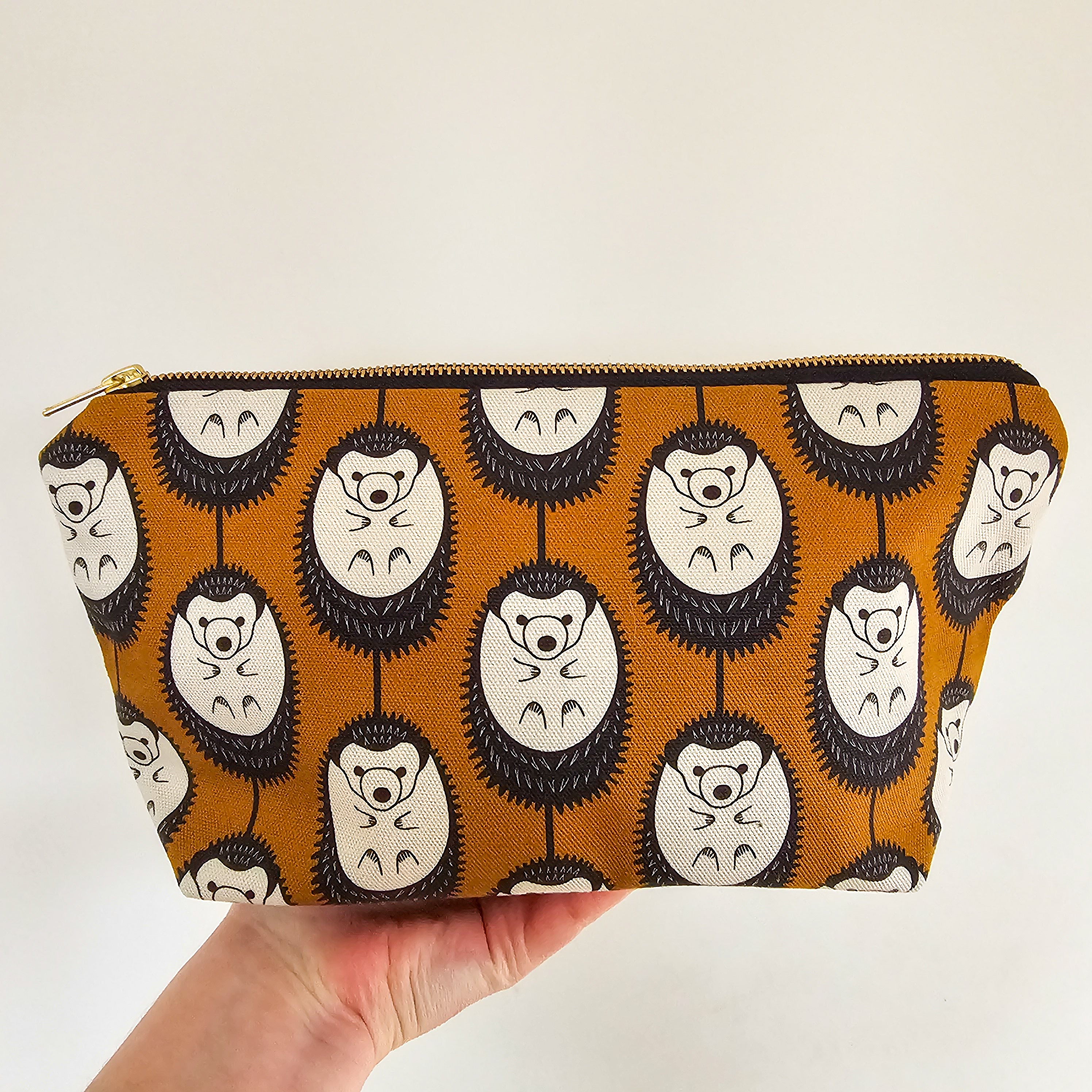 Organic cotton hedgehog zip bag – handmade in the UK by Hannah Issi. Brown & white hedgehog print, versatile and sustainable accessory. Hand holding bag with neutral background.
